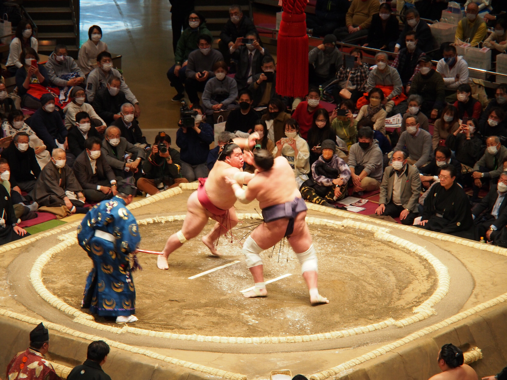 Two sumo wrestlers clash in the center of the ring as the judge and crowd watch intently.